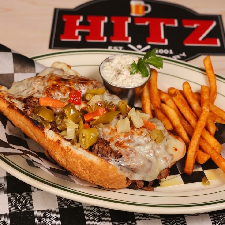 Cheesesteak hoagie topped with melted cheese, peppers, and onions, served with fries and coleslaw on a diner plate.