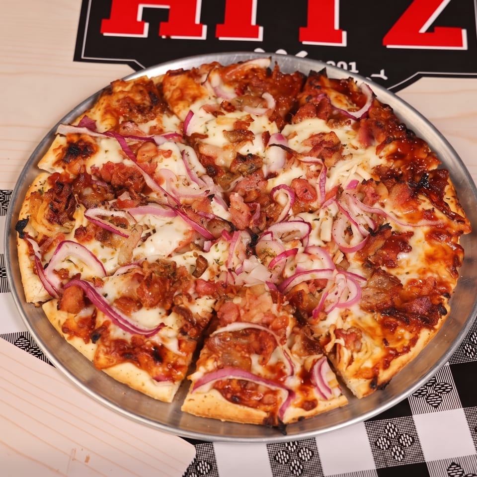 Loaded square pizza topped with melted cheese, meats, and red onion on a metal tray over a patterned tablecloth.