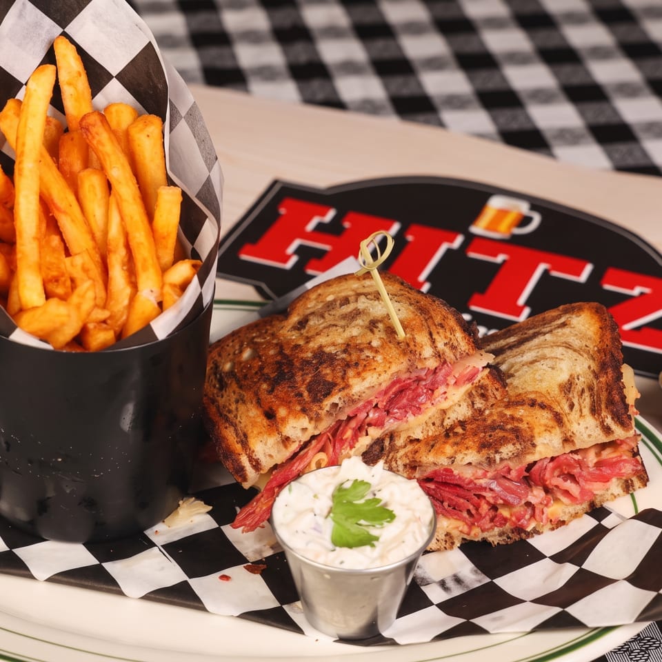 Grilled pastrami sandwich with fries, creamy dipping sauces, and checkerboard paper on a restaurant plate.